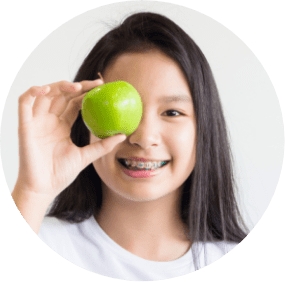 Young girl holding apple up to her eye smiling wearing braces